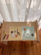 A small wooden children's book stand on a wooden floor in front of sheer white curtains. The stand holds an open children's book with two illustrated pages and two small colored pencils.