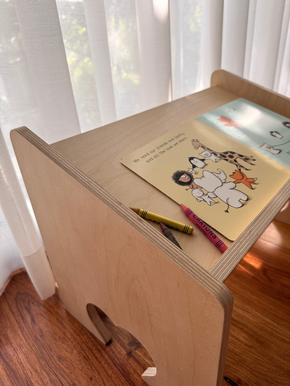 A light natural wood children's desk with a rounded corner and a small cutout, styled with a children's book and crayons on top, set in a room with sheer white curtains and a wooden floor.