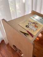 A light natural wood children's desk with a rounded corner and a small cutout, styled with a children's book and crayons on top, set in a room with sheer white curtains and a wooden floor.