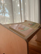 A children's picture book with illustrated pages is propped open on a light wood bookshelf or desk next to window blinds.