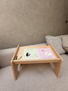 A light natural wood folding lap desk or tray table set up on a textured gray sofa. The desk holds a pastel-colored children's activity book with cartoon illustrations and a small yellow pencil.