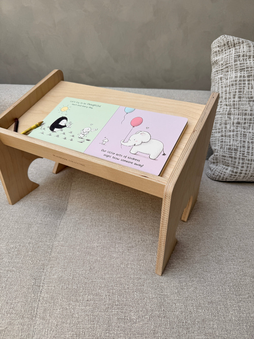A light natural wood toddler lap desk with a slanted writing tray, shown on a textured gray sofa. The tray holds a pastel coloring book with a penguin and an elephant illustration, along with a small yellow pencil.