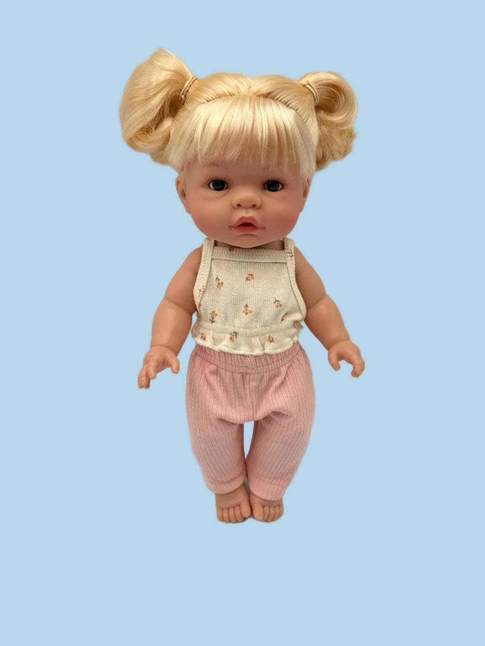 A baby doll with blonde hair in pigtails, wearing a cream sleeveless top with small yellow floral prints and pink pants, standing against a light gray background.