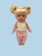 A baby doll with blonde hair in pigtails, wearing a cream sleeveless top with small yellow floral prints and pink pants, standing against a light gray background.