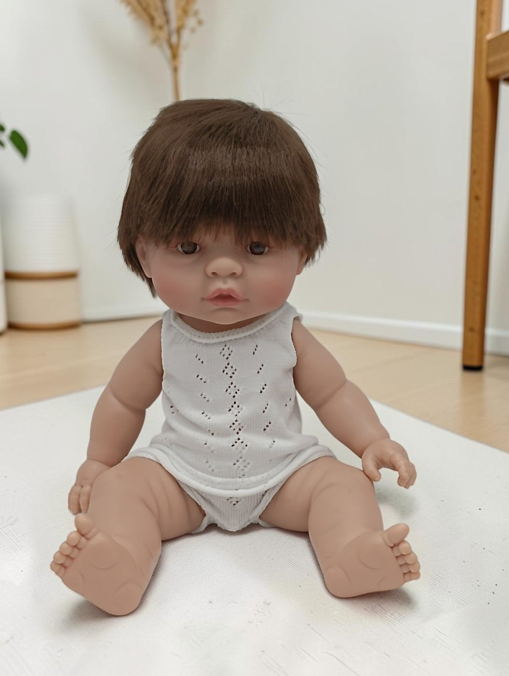 Realistic baby doll with brown hair wearing a white romper on a light surface.