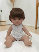 Realistic baby doll with brown hair wearing a white romper on a light surface.