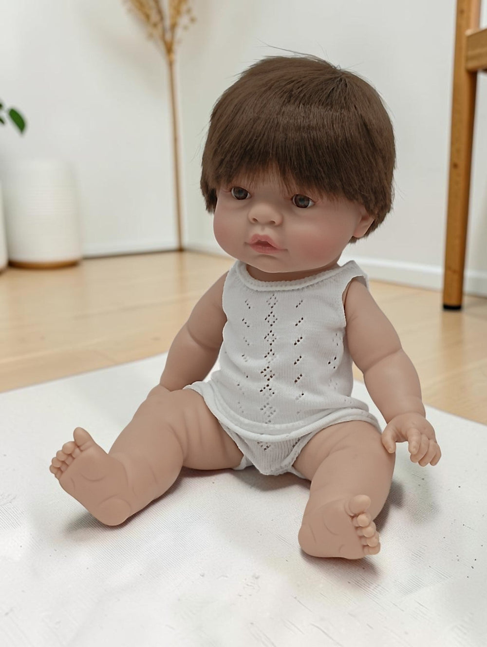 Realistic baby doll with brown hair wearing a white sleeveless top and diaper on a light surface.