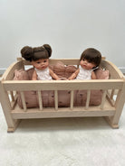 A small wooden doll crib with pink cushioning, holding two baby dolls dressed in white outfits, set against a plain light gray wall and floor.