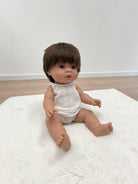 A realistic baby doll with brown hair and skin sits on a white textured rug in a room with light wood flooring and a white wall.
