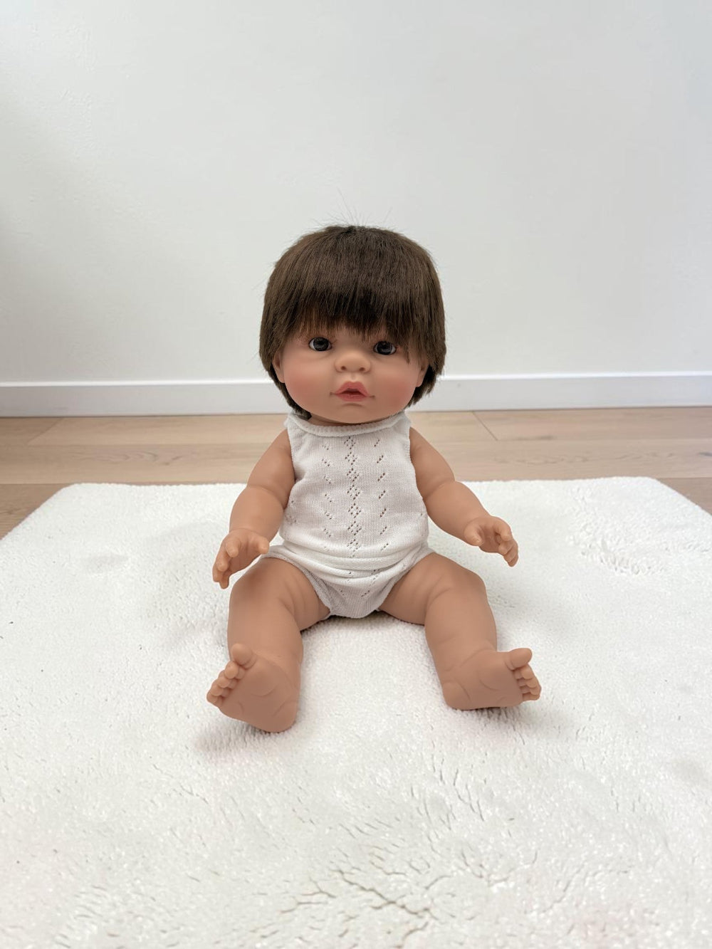 A realistic baby doll with short brown hair sits on a white textured rug in a room with light wood flooring and a white wall.