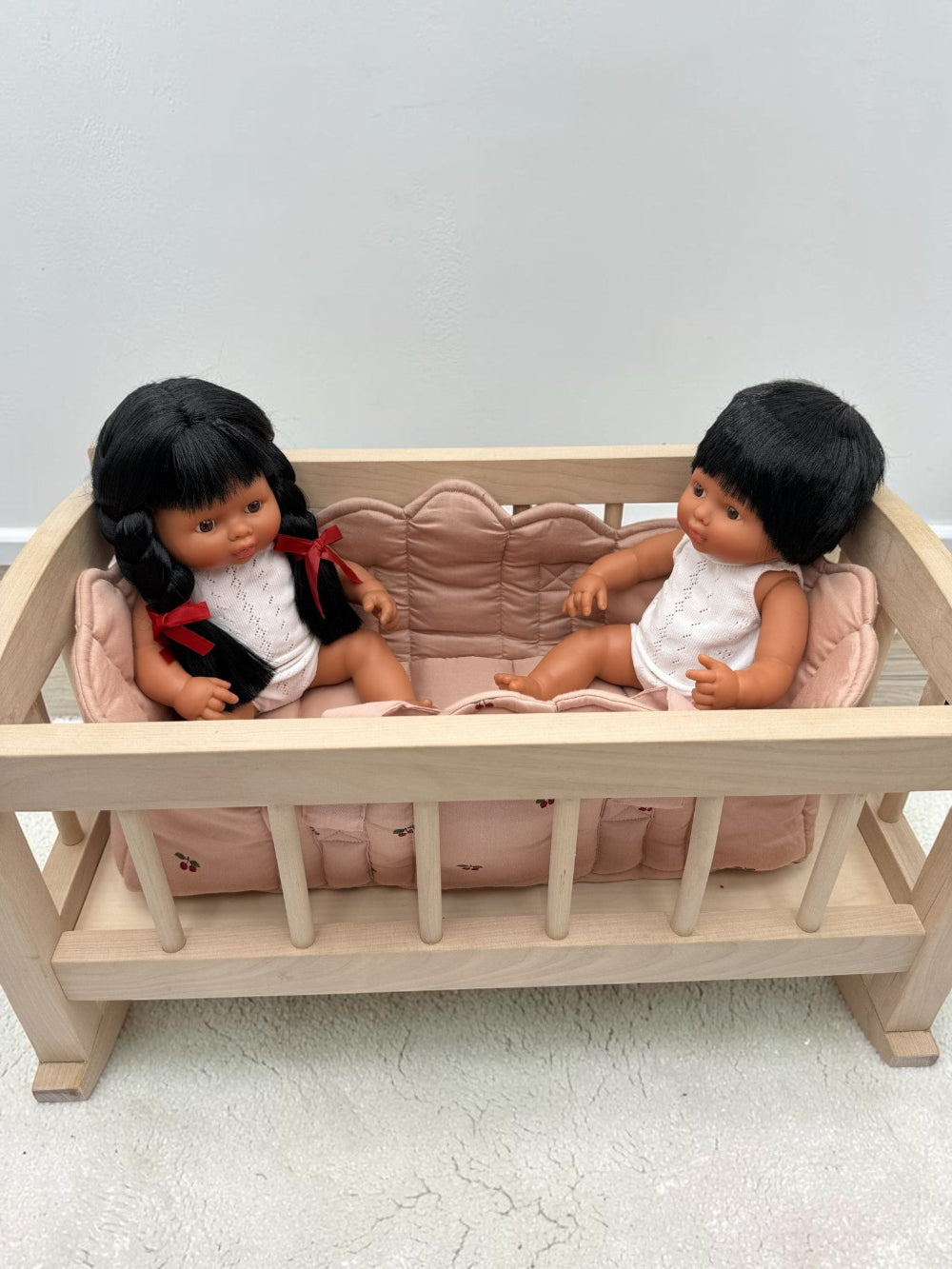 A wooden baby crib with a pink cushion, holding two baby dolls with black hair, one wearing a red and white outfit and the other in a white outfit, set against a plain light gray wall and concrete floor.