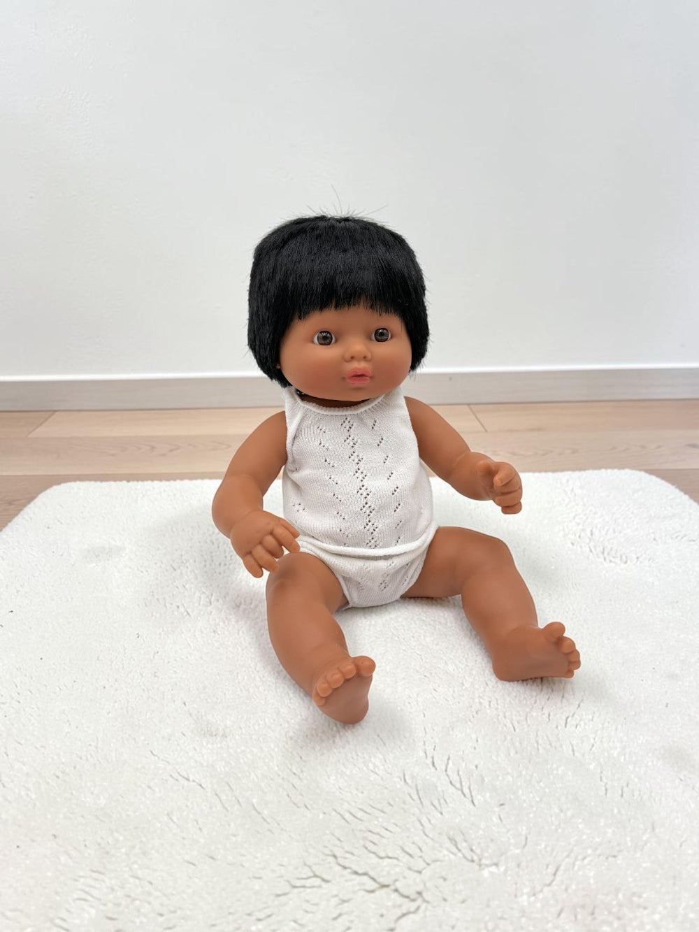 A baby doll with black hair sitting on a white textured rug in front of a white wall.