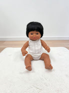 A baby doll with black hair and brown skin sitting on a white textured rug against a white wall and light wood floor.