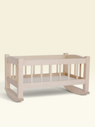 A light natural wood baby rocking cradle with vertical slats and a white mattress, shown against a solid pale cream background.