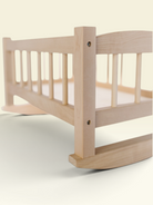 A close-up of a light natural wood rocking bed frame, showing the slatted side rail, corner post, and curved rocker base against a soft cream background.