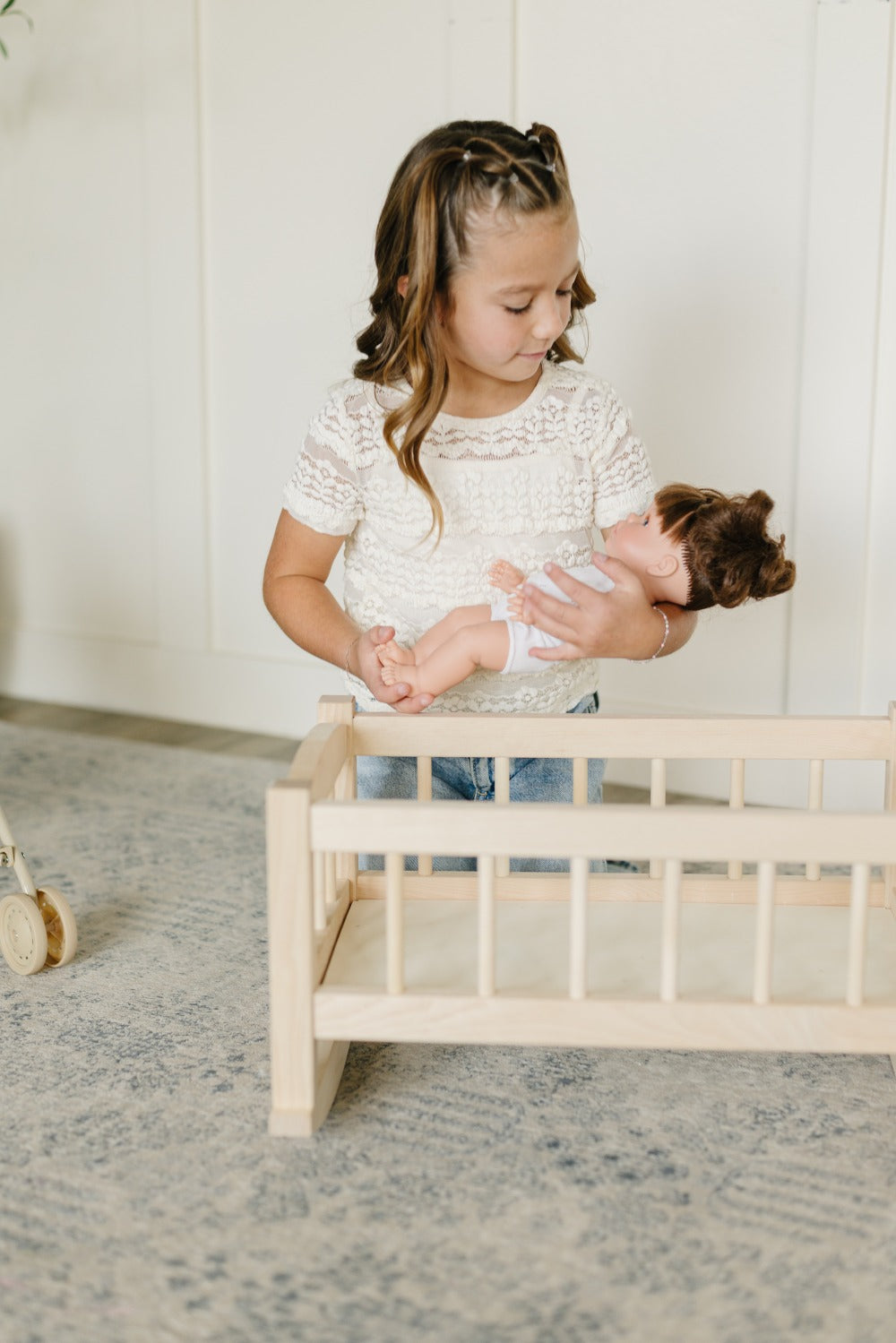 A young girl with braided hair wearing a white lace top and blue jeans holds a doll next to a small light wood crib in a bright room with a patterned rug.