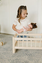 A young girl with braided hair wearing a white lace top and blue jeans holds a doll next to a small light wood crib in a bright room with a patterned rug.