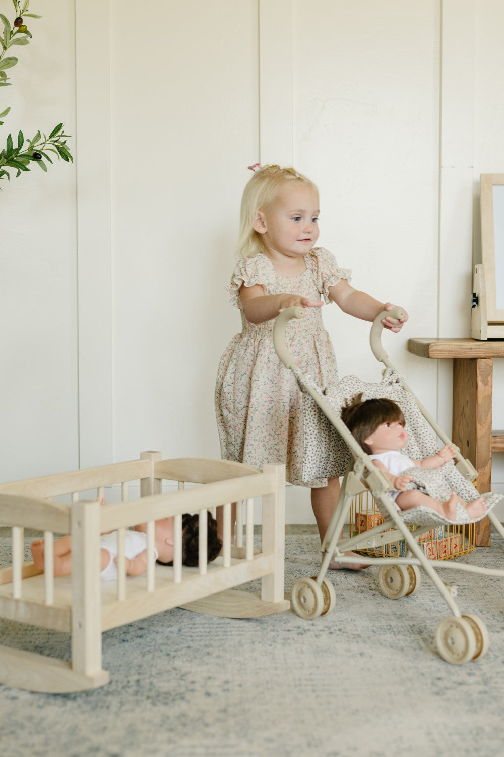 A young toddler girl in a beige patterned dress pushes a light wood toy stroller with a doll inside, next to a wooden rocking cradle with another doll, in a bright, neutral room.