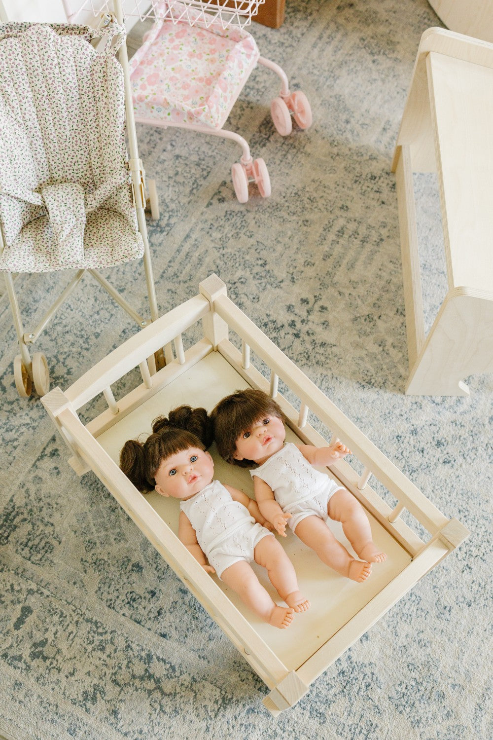 Two baby dolls lying on a cream fabric mattress inside a light wood bassinet, placed on a blue-gray patterned rug in a nursery setting with a stroller and changing table visible in the background.