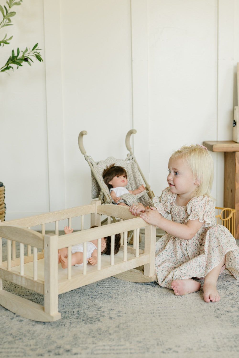 A young blonde toddler in a cream floral dress sits on a gray rug next to a light wood doll crib, playing with a doll in a matching woven rocking chair. The scene is set in a bright, neutral nursery with white paneled walls.