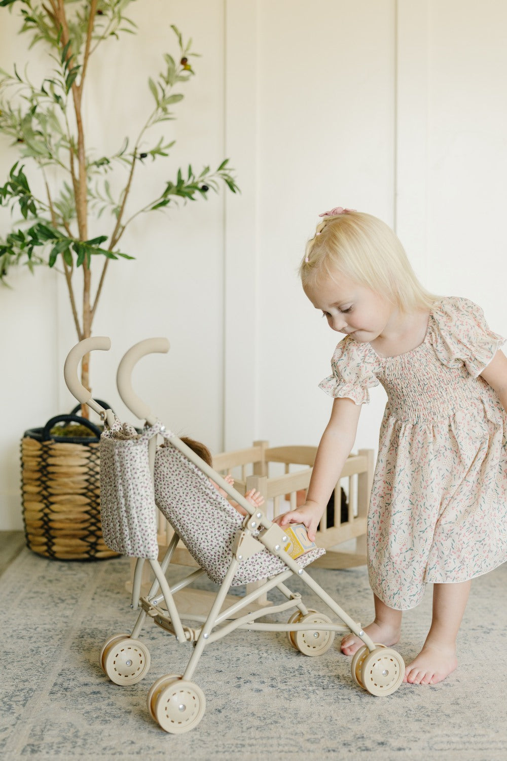 A young blonde girl in a light floral dress pushes a wooden toy stroller with a patterned fabric canopy in a bright, neutral room.