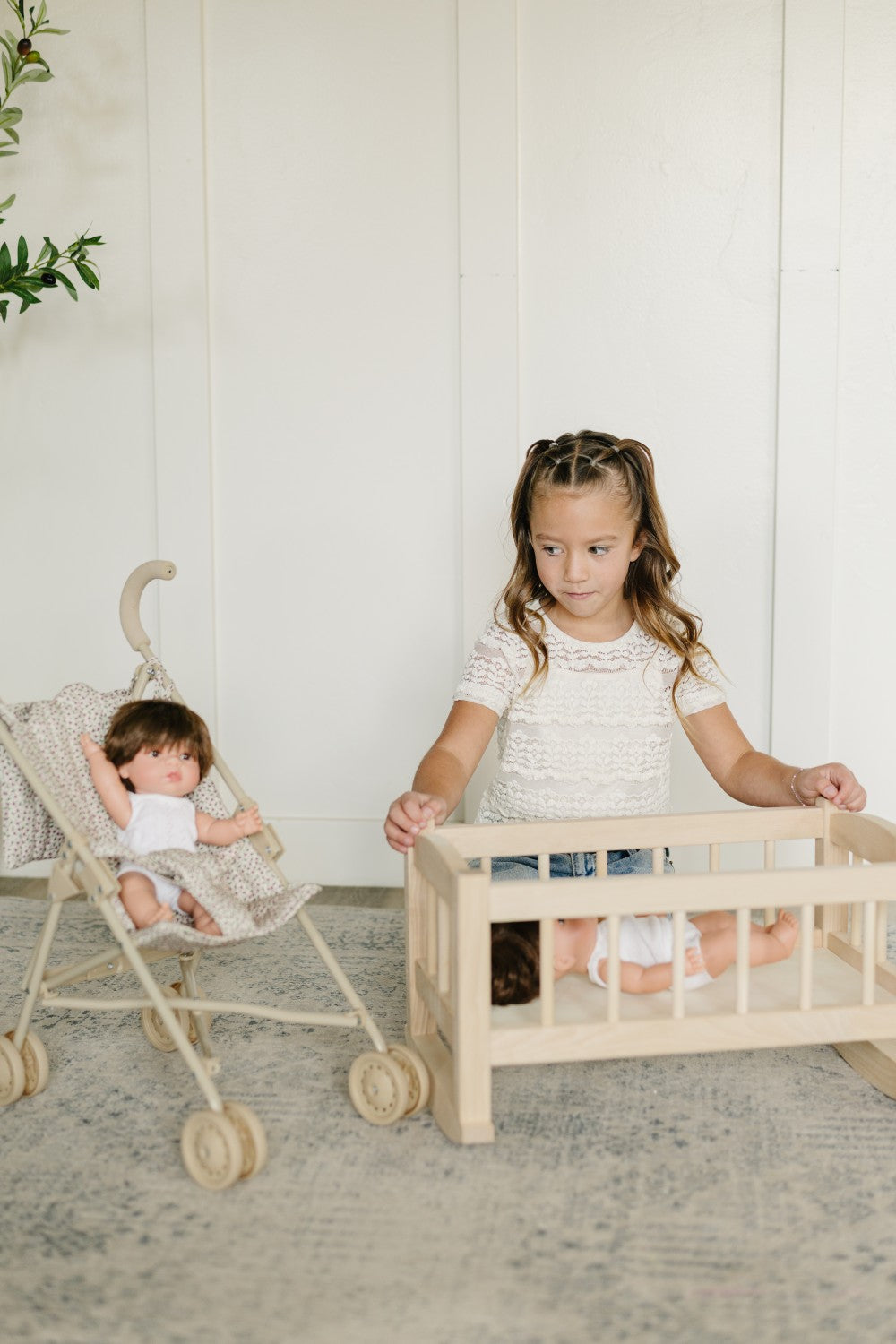 A young girl stands behind a light wood doll crib while a baby doll sits inside, with a stroller holding another doll to the left, set against a white paneled wall and a textured rug.