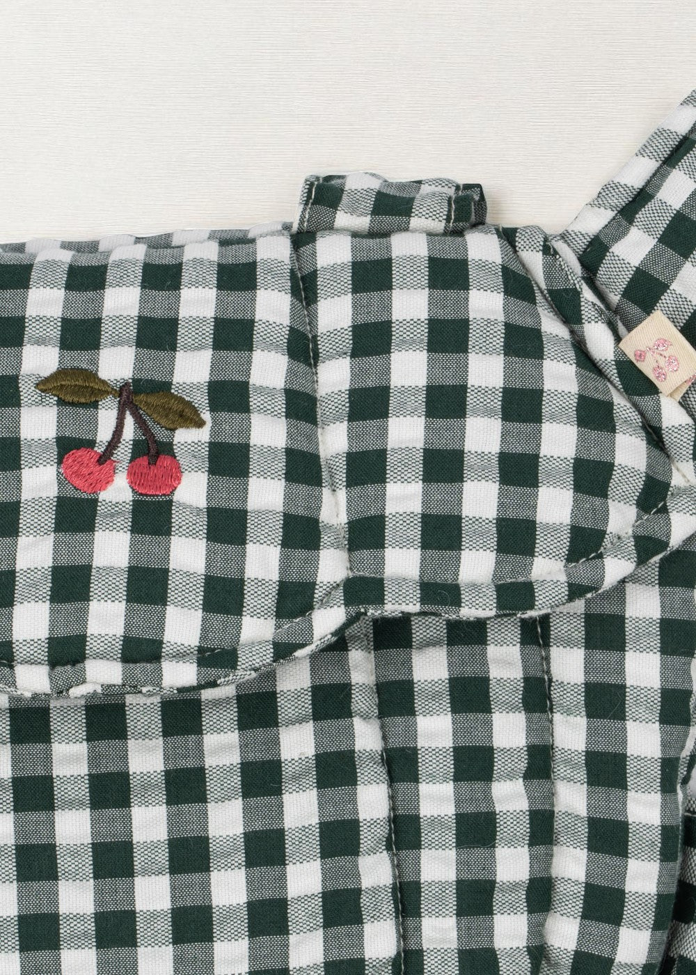 Close-up of embroidered cherry accent on green and white checkered doll carrier showing whimsical farmhouse detail