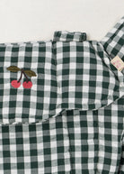 Close-up of embroidered cherry accent on green and white checkered doll carrier showing whimsical farmhouse detail