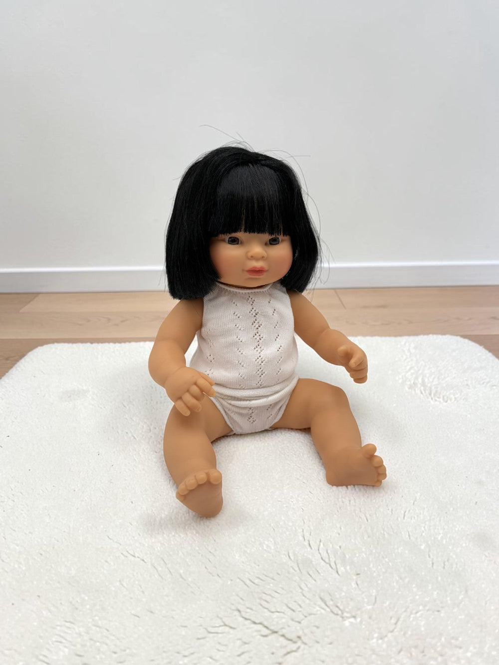 A baby doll with black hair sitting on a white textured rug against a plain white wall and light wood floor.