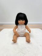 A realistic baby doll with black hair sitting on a white textured rug in front of a plain wall.