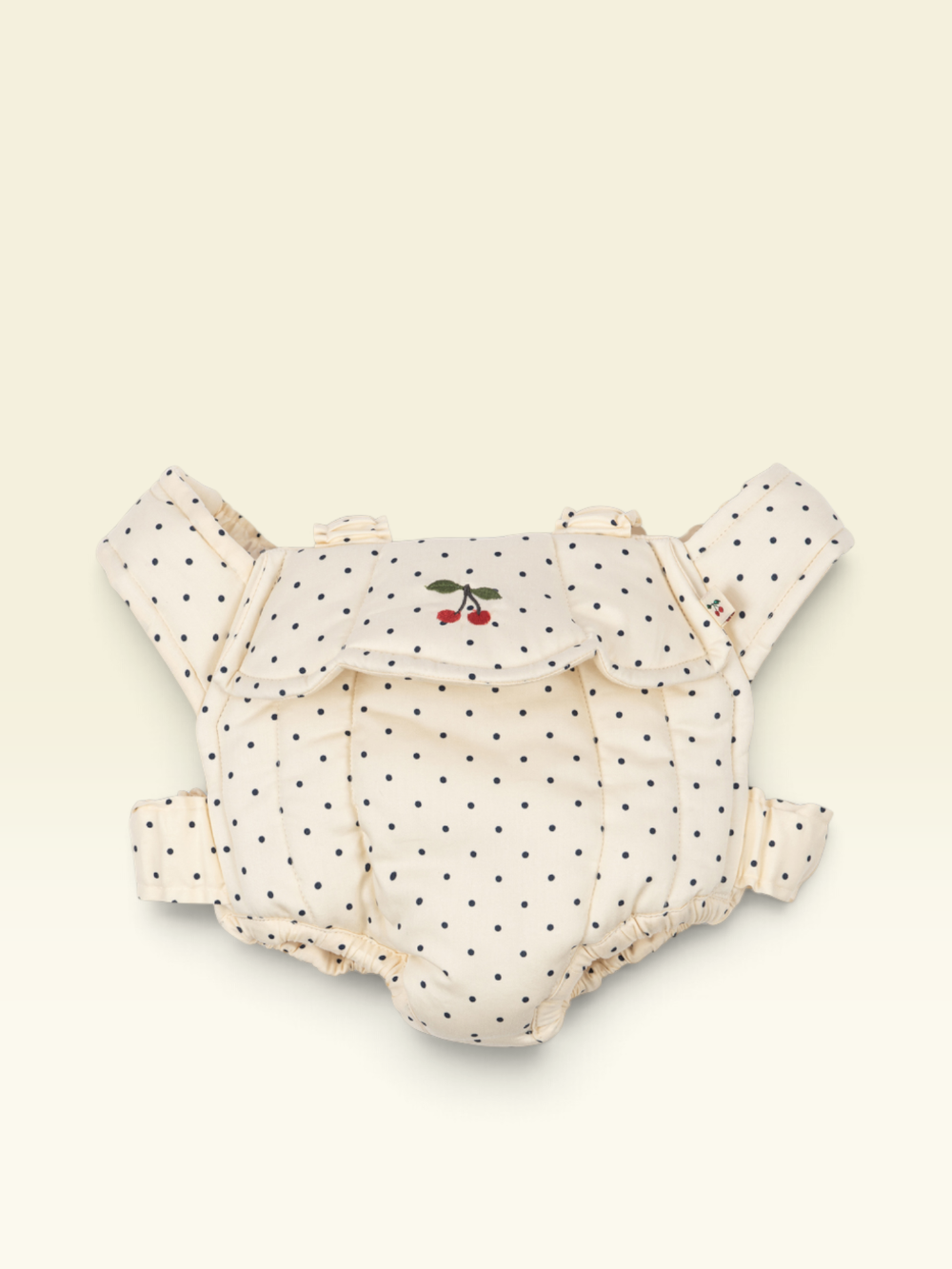 A cream-colored baby carrier with black polka dots and a small embroidered cherry motif on the front flap, displayed against a solid cream background.