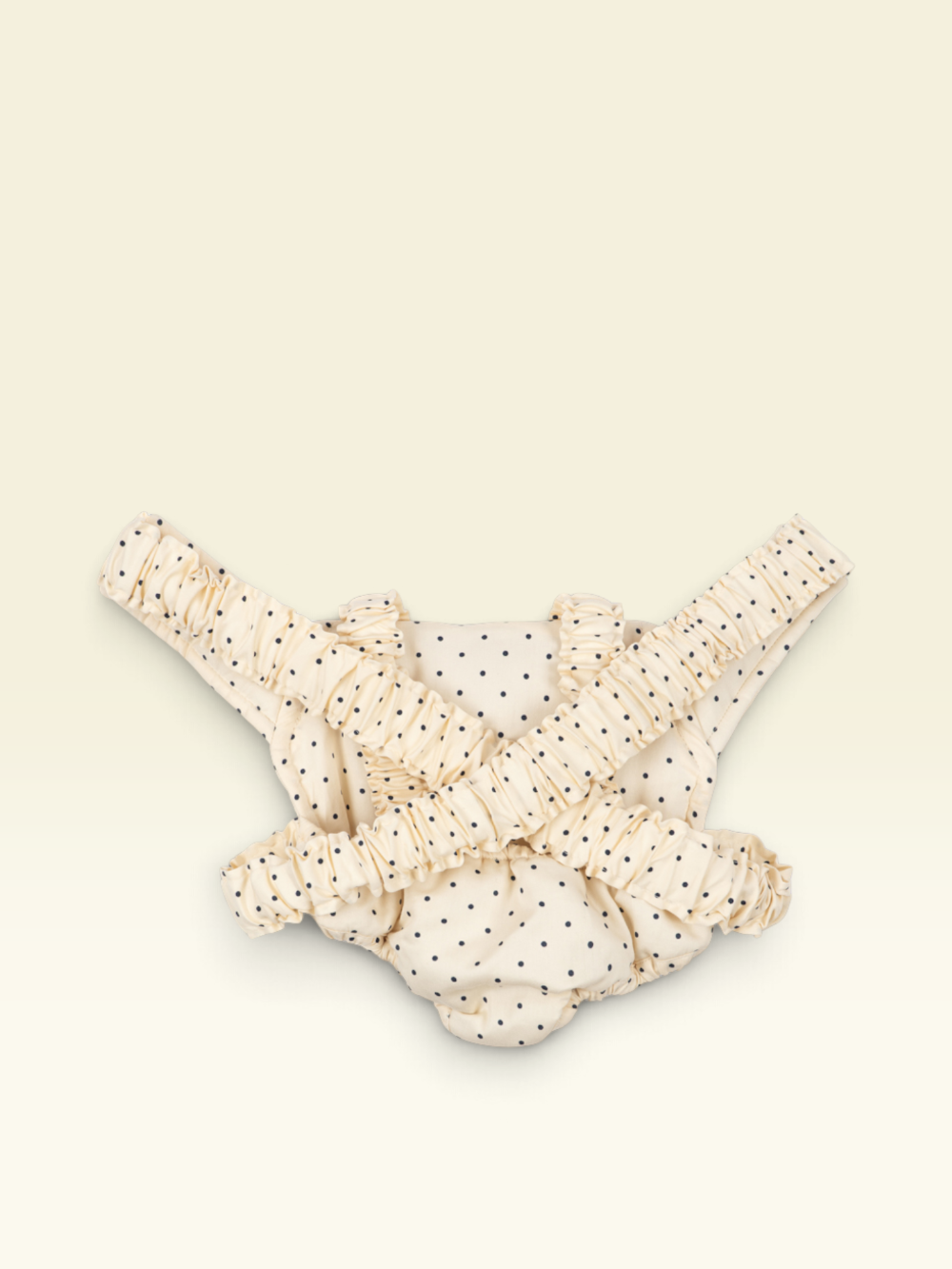 A cream-colored baby harness with black polka dots and ruffled straps, laid flat on a light cream background.
