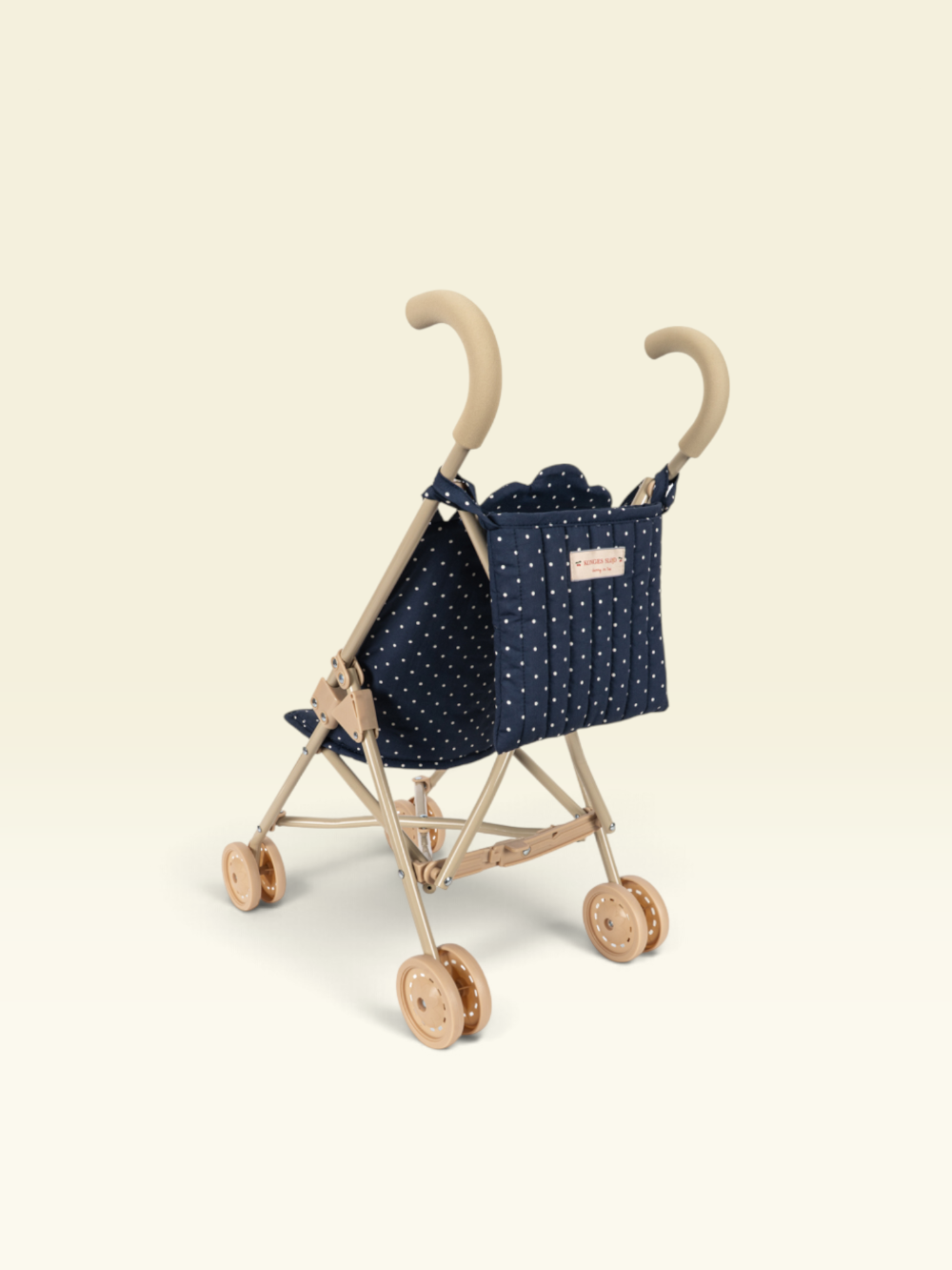 A compact stroller with a natural wood frame, beige handles, and a navy blue fabric seat cover decorated with small white polka dots. The stroller is photographed from a high-angle perspective against a solid, warm off-white background.