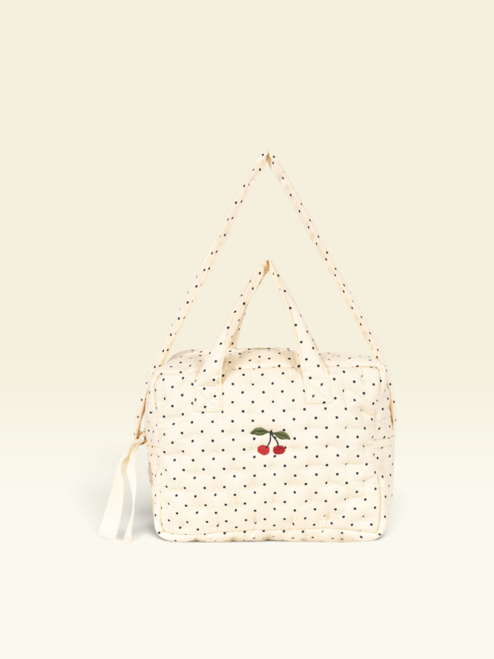 A cream-colored quilted bag with black polka dots and a small embroidered cherry motif on the front, featuring a matching shoulder strap and top handles, set against a solid cream background.