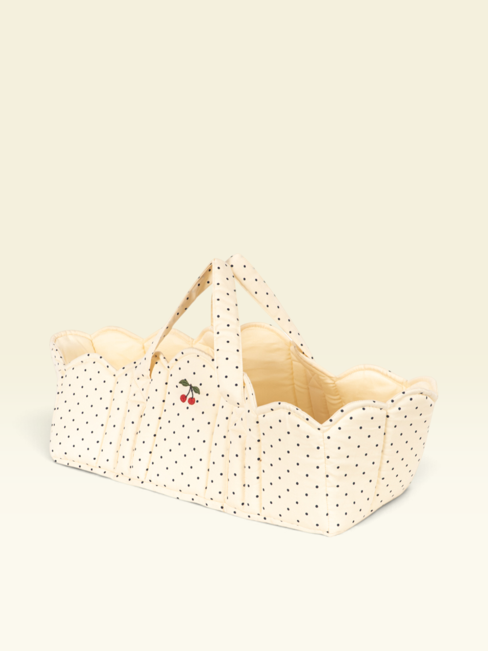 A cream-colored fabric bassinet or basket with black polka dots, featuring a small cherry embroidery on the front and matching fabric handles, set against a solid cream background.