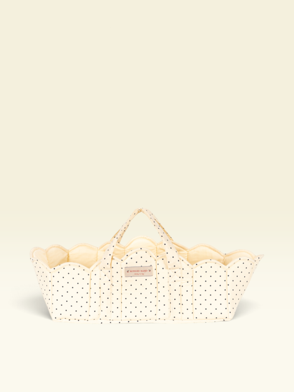 A cream-colored fabric basket with black polka dots and scalloped edges, featuring two top handles and a small pink brand label, set against a solid cream background.