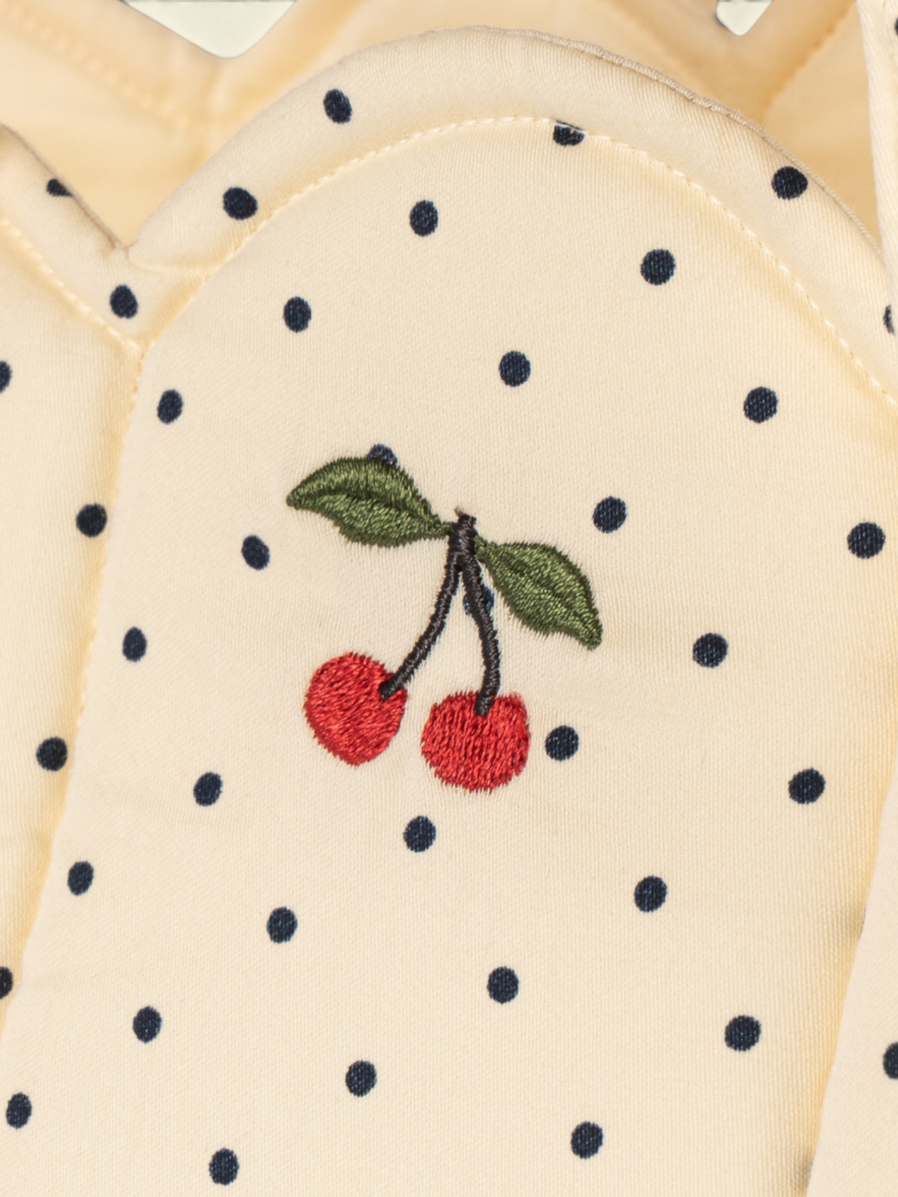 Close-up of a cream-colored fabric garment with black polka dots and an embroidered cherry motif in red, green, and black.