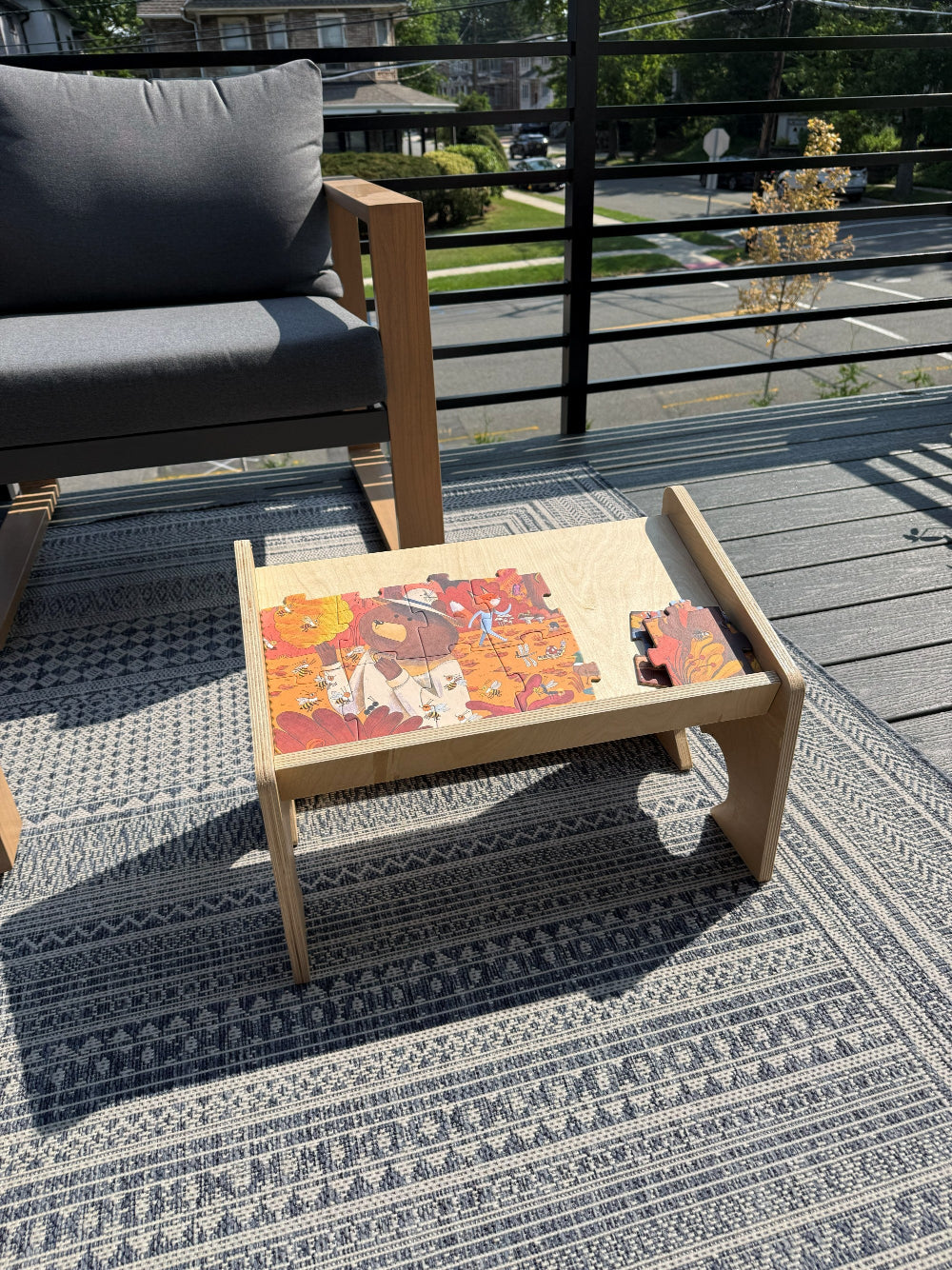 A light natural wood low coffee table with a slanted tray top, styled on a patterned outdoor rug on a wooden deck. The tray contains a colorful jigsaw puzzle and a small red toy.