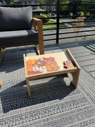 A light natural wood low coffee table with a slanted tray top, styled on a patterned outdoor rug on a wooden deck. The tray contains a colorful jigsaw puzzle and a small red toy.