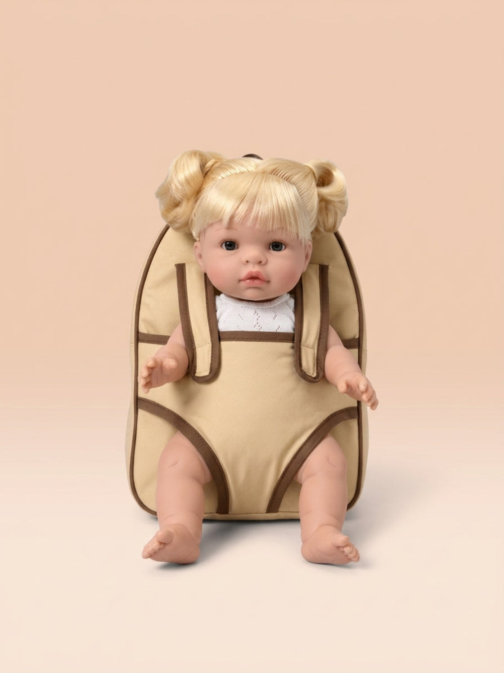 A blonde doll with pigtails sits inside a beige baby carrier or backpack-style seat with brown trim, photographed against a soft peach studio background.