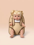 A blonde doll with pigtails sits inside a beige baby carrier or backpack-style seat with brown trim, photographed against a soft peach studio background.