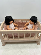 A light natural wood doll bed with a pink cushion, containing two small dolls with black hair and white outfits, set against a plain light gray wall and textured floor.