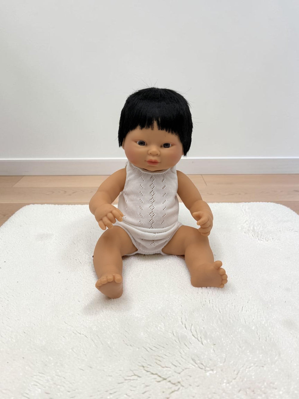 A baby doll with black hair wearing a white sleeveless top and white diaper cover, sitting on a white textured rug against a plain white wall and light wood floor.