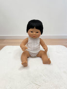 A baby doll with black hair wearing a white sleeveless top and white diaper cover, sitting on a white textured rug against a plain white wall and light wood floor.