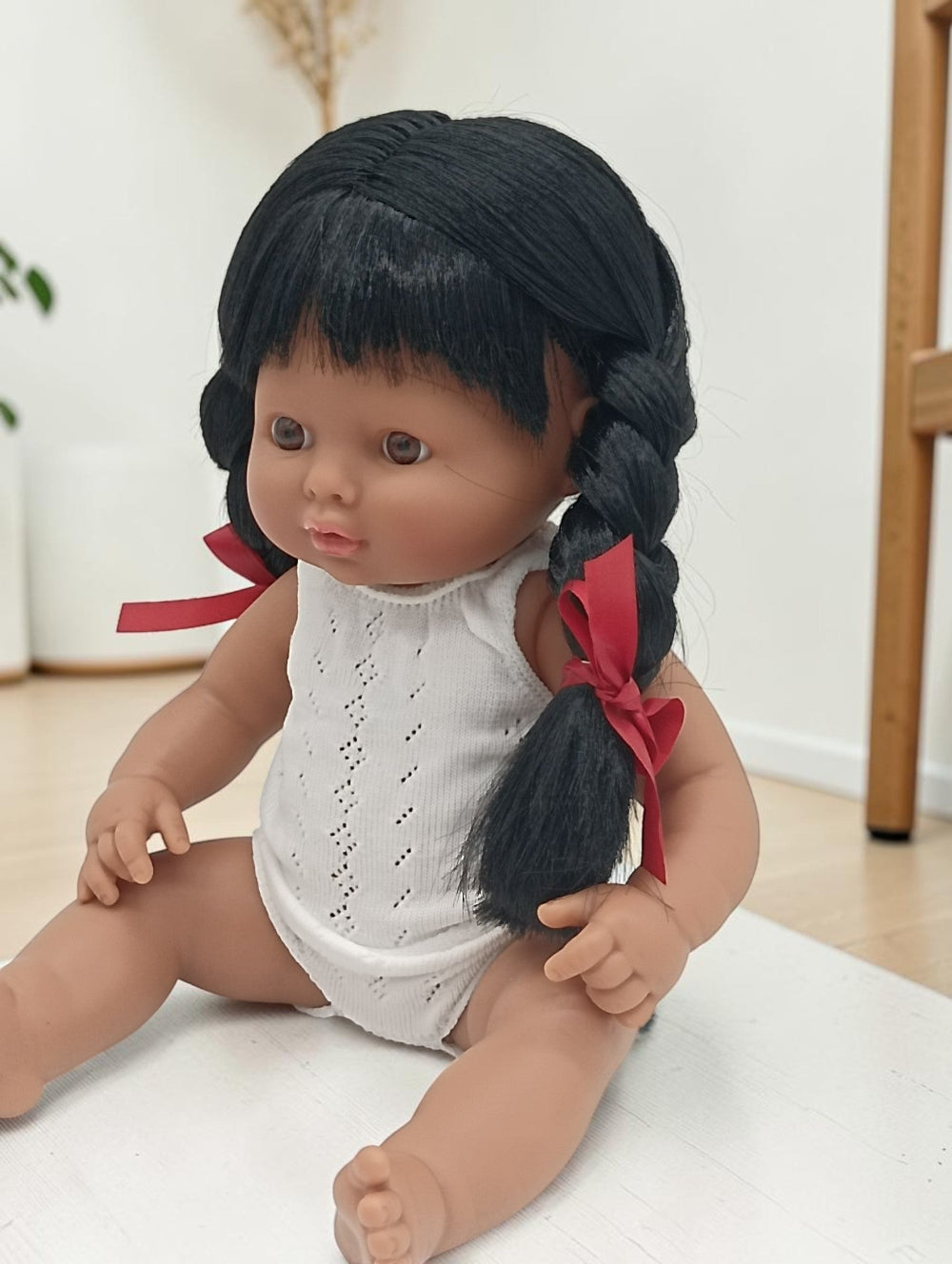 Doll with braided black hair and red ribbons wearing a white outfit on a light surface.