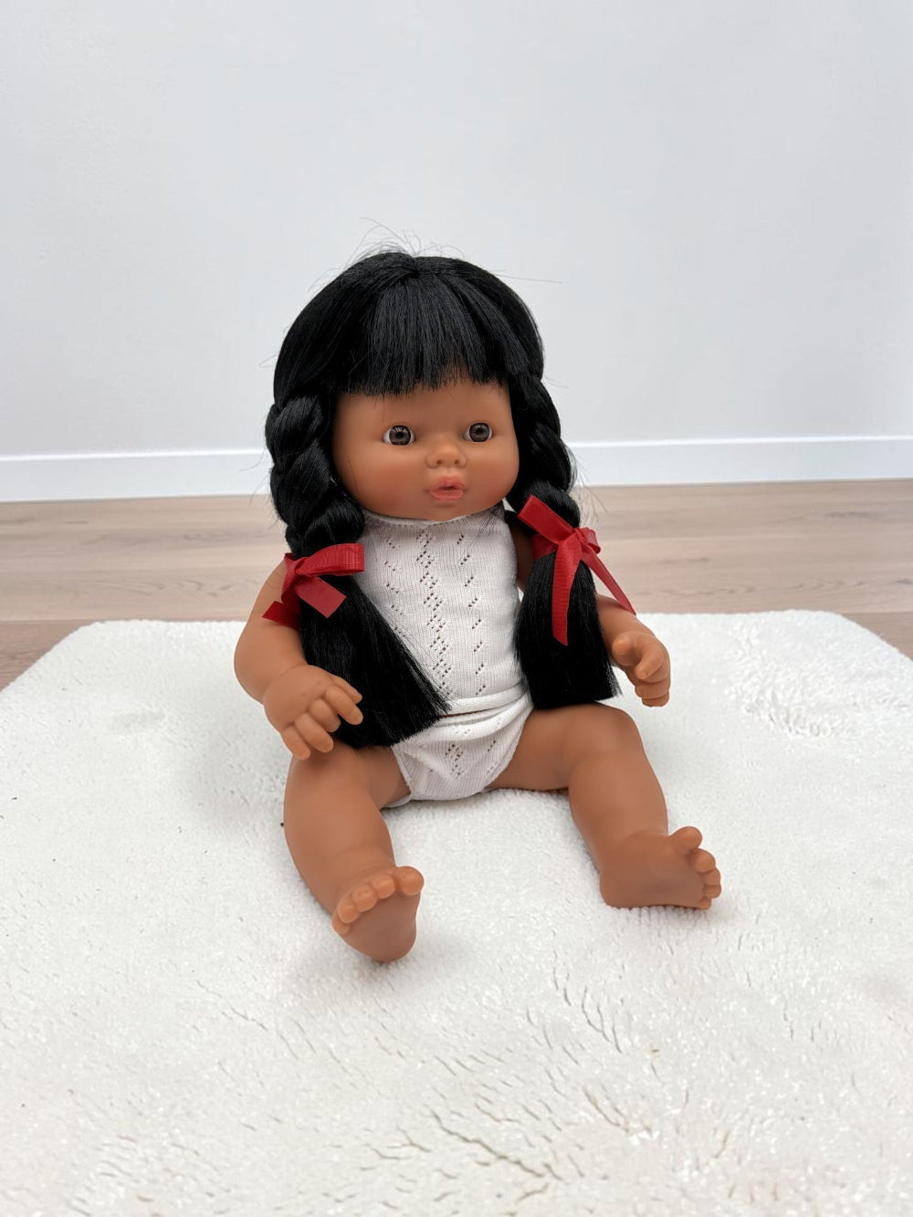 A baby doll with black braided hair and red ribbon accents, wearing a white knit top and white diaper, sitting on a white textured rug.