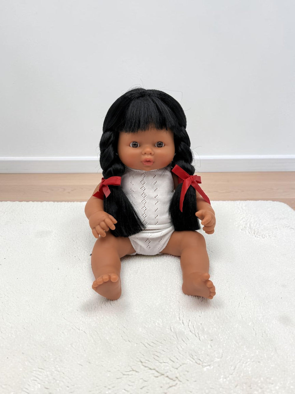 A realistic baby doll with dark brown skin, black braided hair, and a white knit top with red ribbon accents, sitting on a white textured rug.