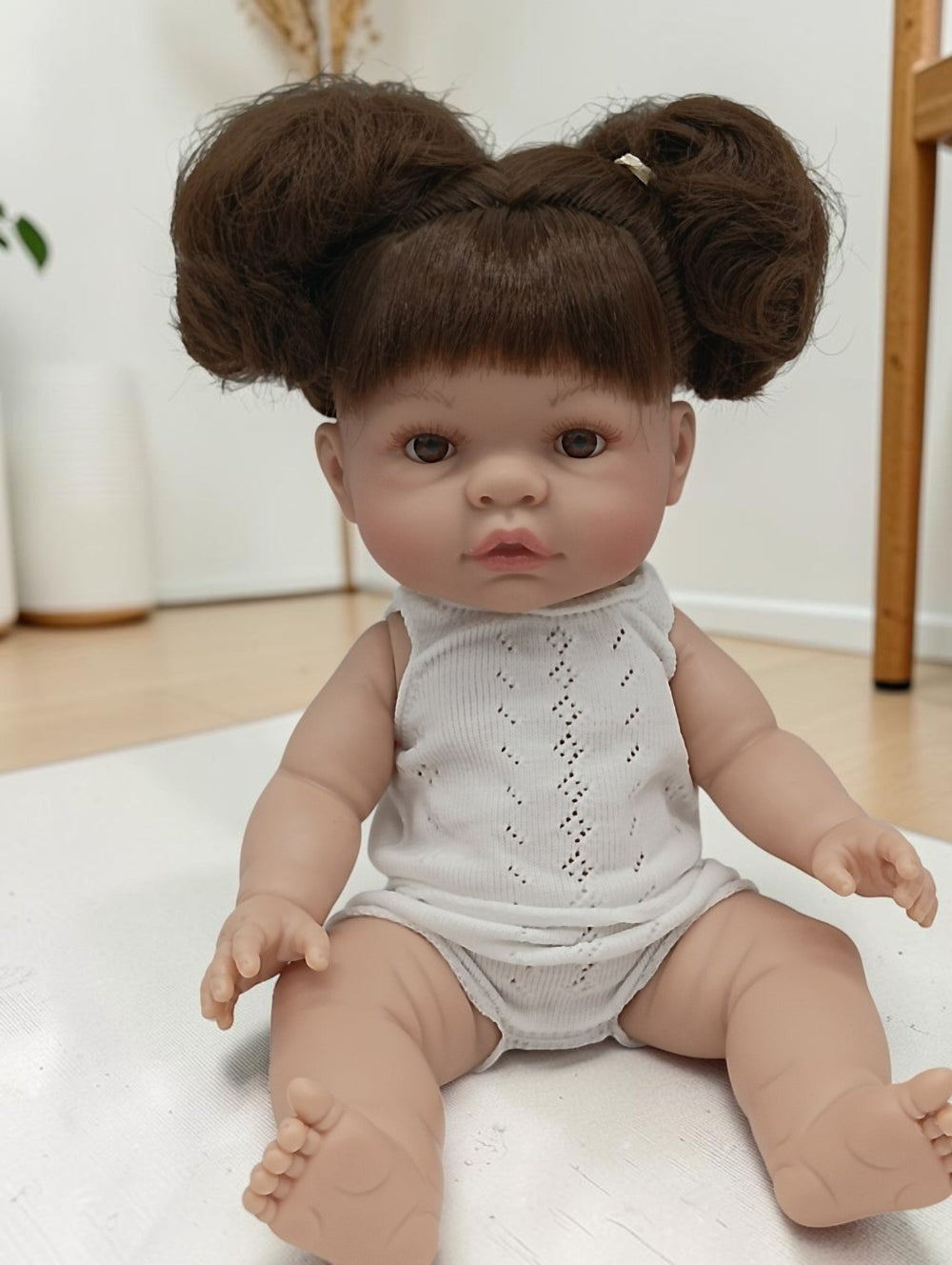 Realistic baby doll with brown hair styled in buns wearing a light gray romper on a white surface.
