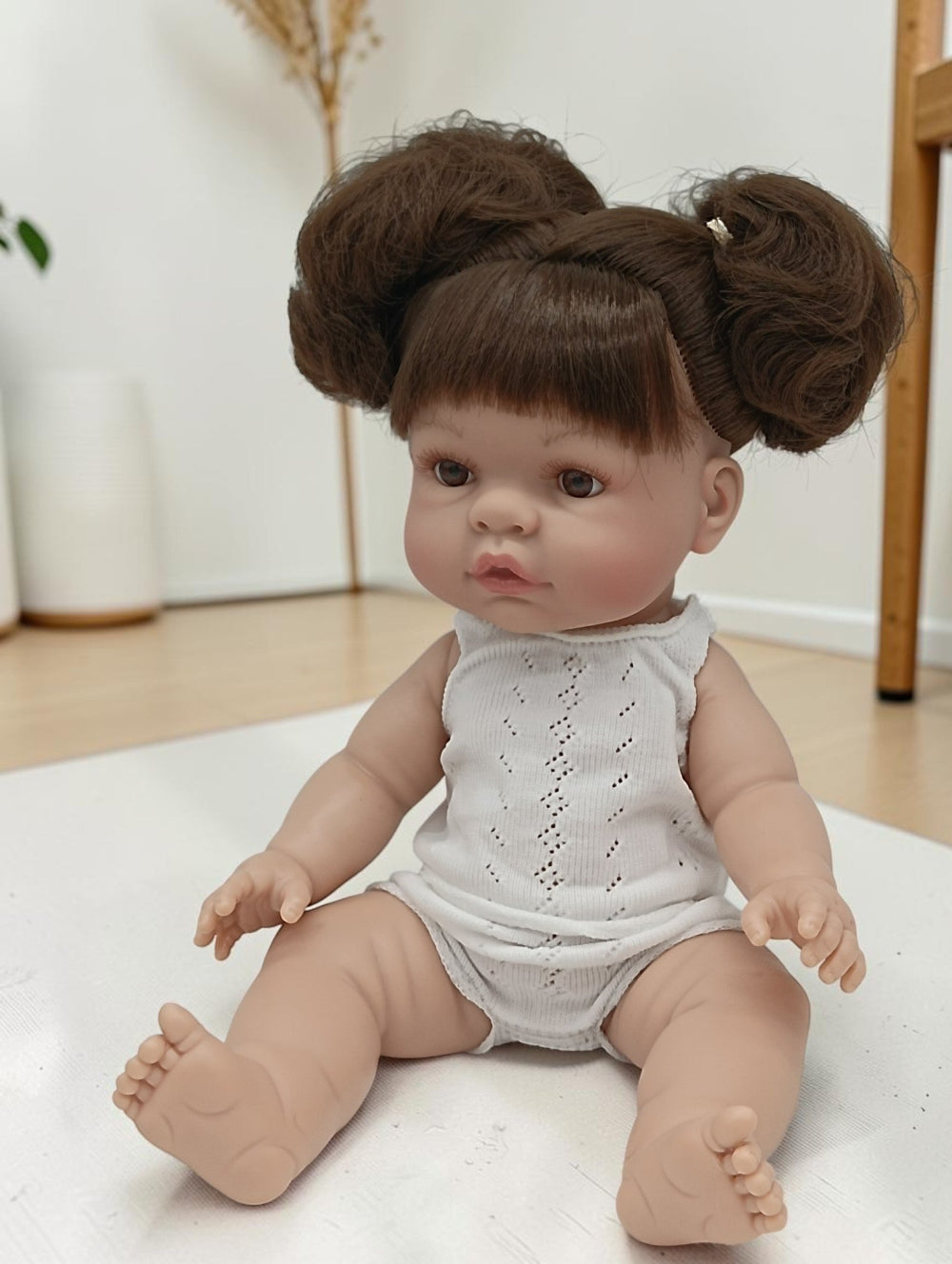 Baby doll with brown hair styled in buns wearing a white sleeveless outfit on a light surface.