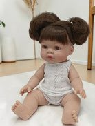 Baby doll with brown hair styled in buns wearing a white sleeveless outfit on a light surface.